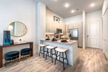 Staged kitchen with breakfast bar, granite countertops, white fabric topped barstools, stainless appliances, taupe cabinetry, circular mirror, wood style flooring - Photo Gallery 33