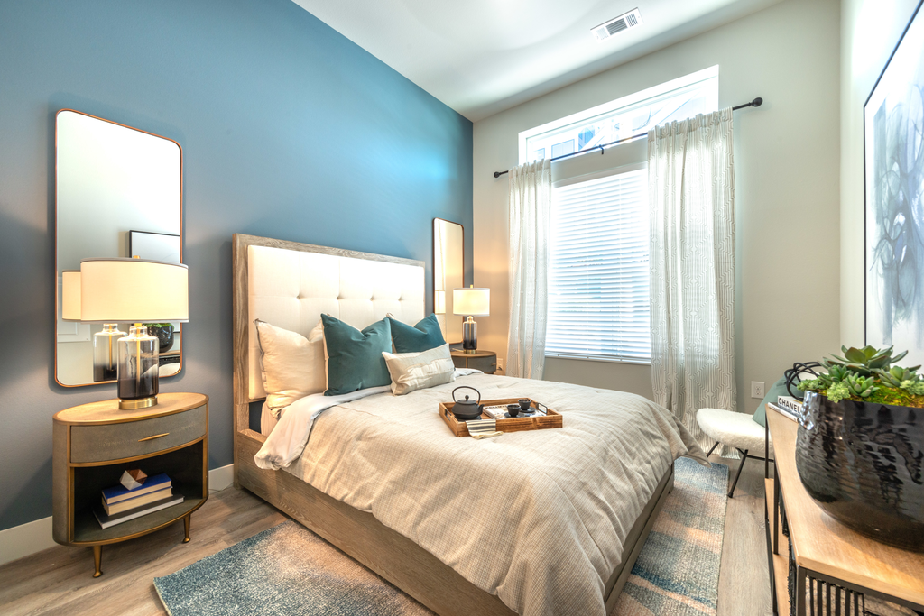 Staged bedroom with bed, accent rug, wood style flooring, oval side tables with lamps and mirrors, large windows with blinds and sheer curtain with blue accent wall