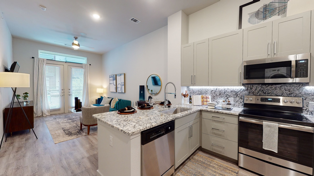 Staged kitchen and livingroom with granite countertops, stainless appliances, taupe cabinetry, wood style flooring, couch, accent rug, windows with curtains, wall art and ceiling fan with light