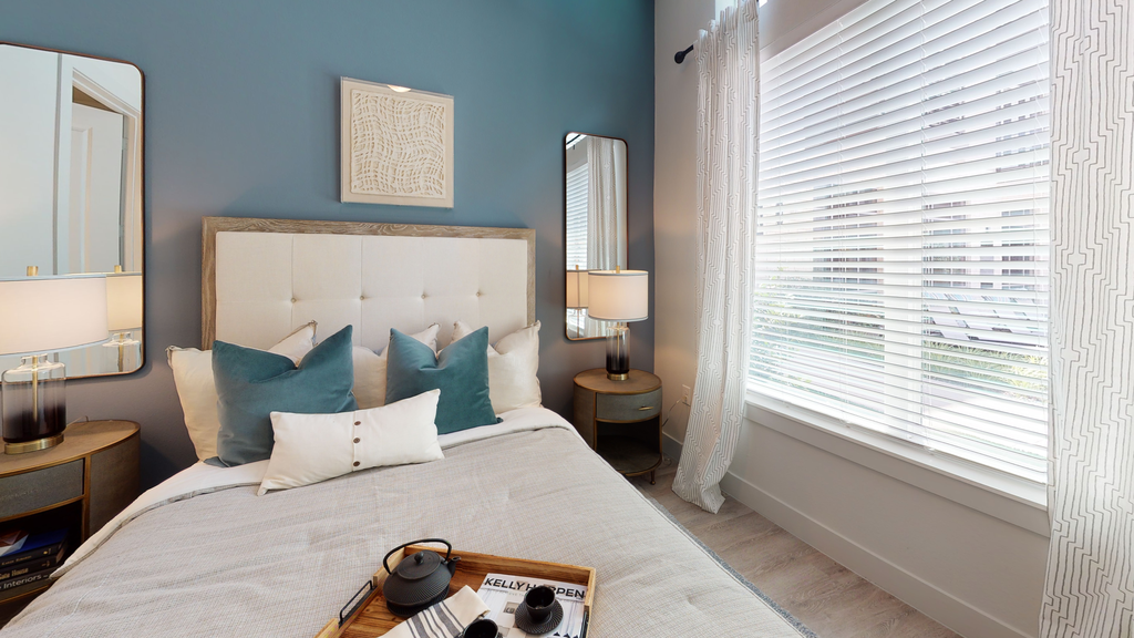 Staged bedroom with bed, accent rug, side tables with lamps and mirrors, and large window with blinds and sheer curtain