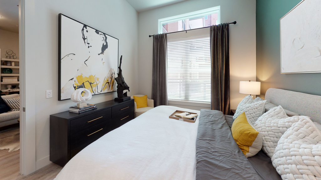 Staged bedroom with bed, dresser, art on wall, accent wall, large window with blinds and curtain
