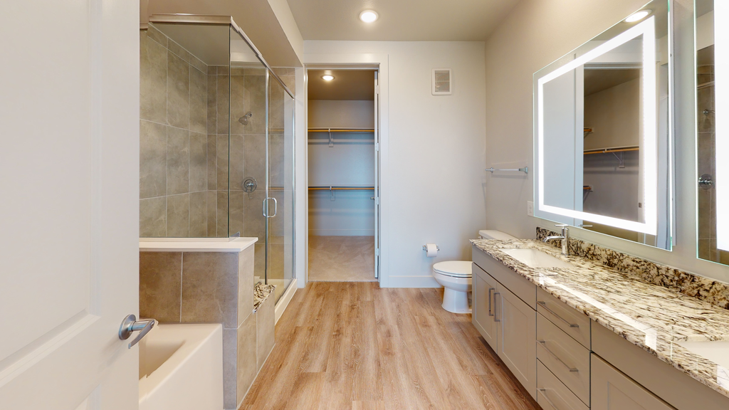 Bathroom with wood style flooring, granite countertops, taupe cabinetry, custom light vanity mirrors, dual vanities, garden tub and walk in shower with direct access to walk in closet