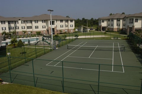 A tennis court surrounded by a green fence and apartment buildings in the background.