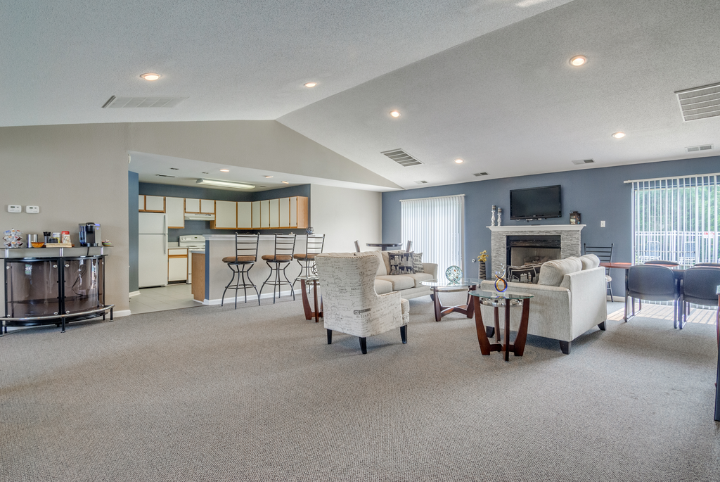 The furnished clubhouse is a large open room with a coffee bar, a full kitchen with breakfast bar, and living area with a fireplace and two sliding glass doors on either side of the fireplace. It is furnished with three bar stools, two couches and an oversized chair, two smaller tables with chairs, and a coffee table.