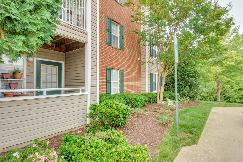 The apartment buildings are brick with tan siding and green shutters. The patios and balconies are enclosed. The building is surrounded by mature trees and lush landscape.