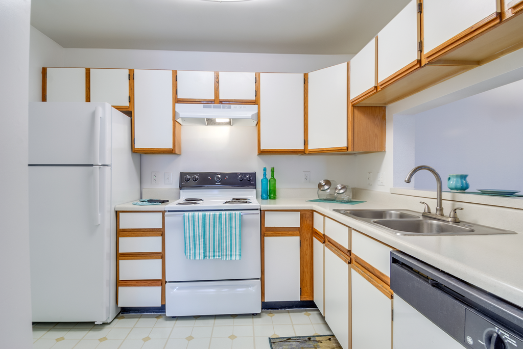The three bedroom kitchen has white cabinets with tan trim, white appliances, formica countertops, and a double basin sink with gooseneck faucet. The breakfast bar has a place setting of fiesta ware for two.