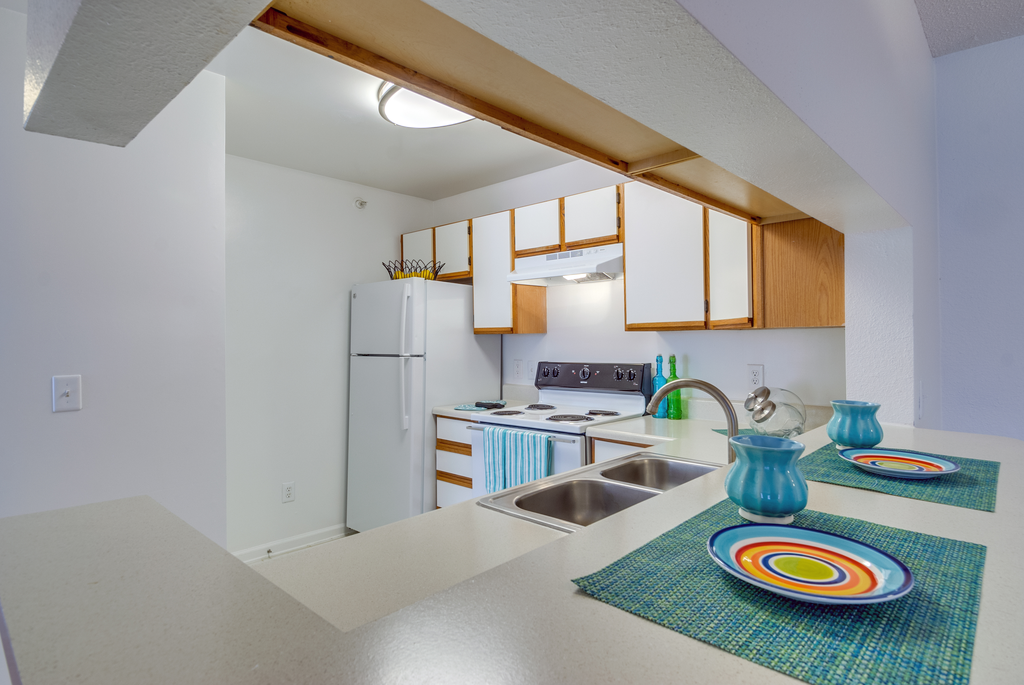 The three bedroom kitchen has white cabinets with tan trim, white appliances, formica countertops, and a double basin sink with gooseneck faucet. The breakfast bar has a place setting of fiesta ware for two.