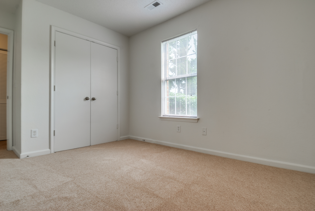 The vacant bedroom has wall to wall carpet, white walls, one standard window, and double doors opening to the closet.