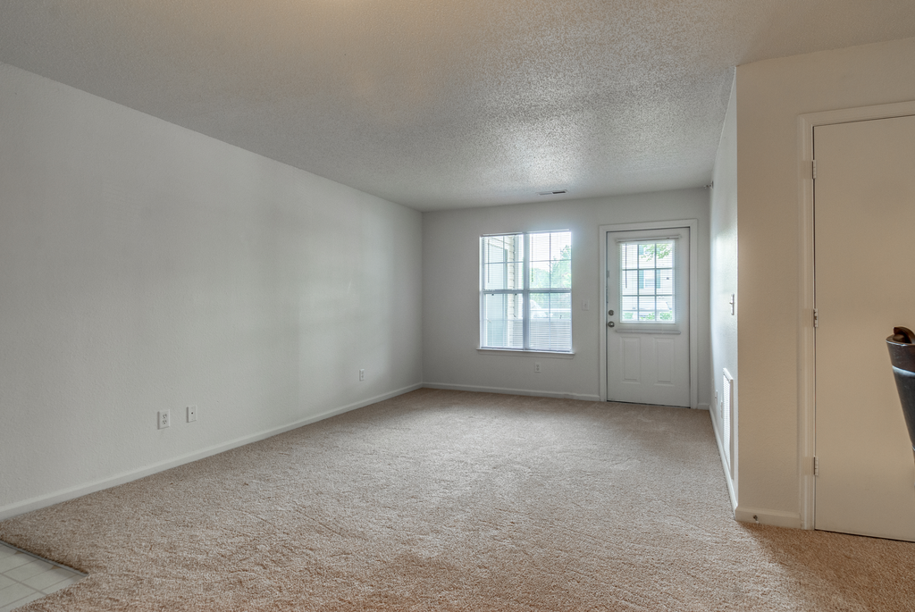 The vacant apartment living room has wall to wall carpet white walls, large picture window and a door leading out to the patio.