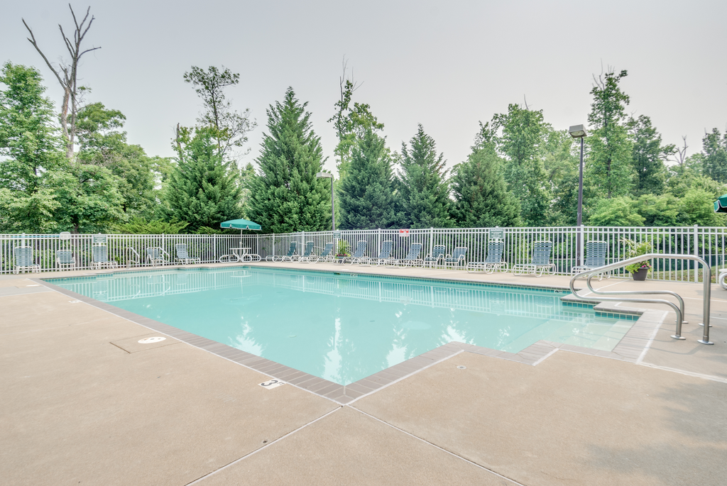 The community pool is rectangular in shape with blue and green pool furniture placed around the sundeck. There is a round picnic table with a green umbrella in the middle. The pool is placed on the back side of the Leasing Office.