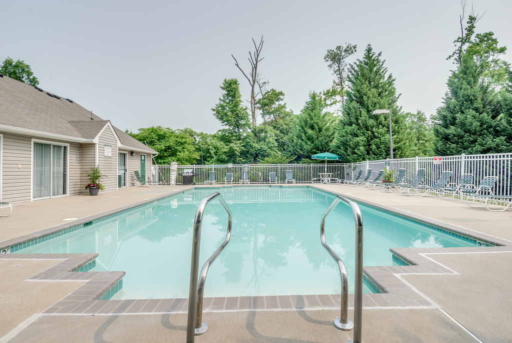 The community pool is rectangular in shape with blue and green pool furniture placed around the sundeck. There is a round picnic table with a green umbrella in the middle. The pool is placed on the back side of the Leasing Office.