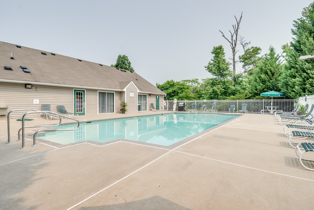 The community pool is rectangular in shape with blue and green pool furniture placed around the sundeck. There is a round picnic table with a green umbrella in the middle. The pool is placed on the back side of the Leasing Office.