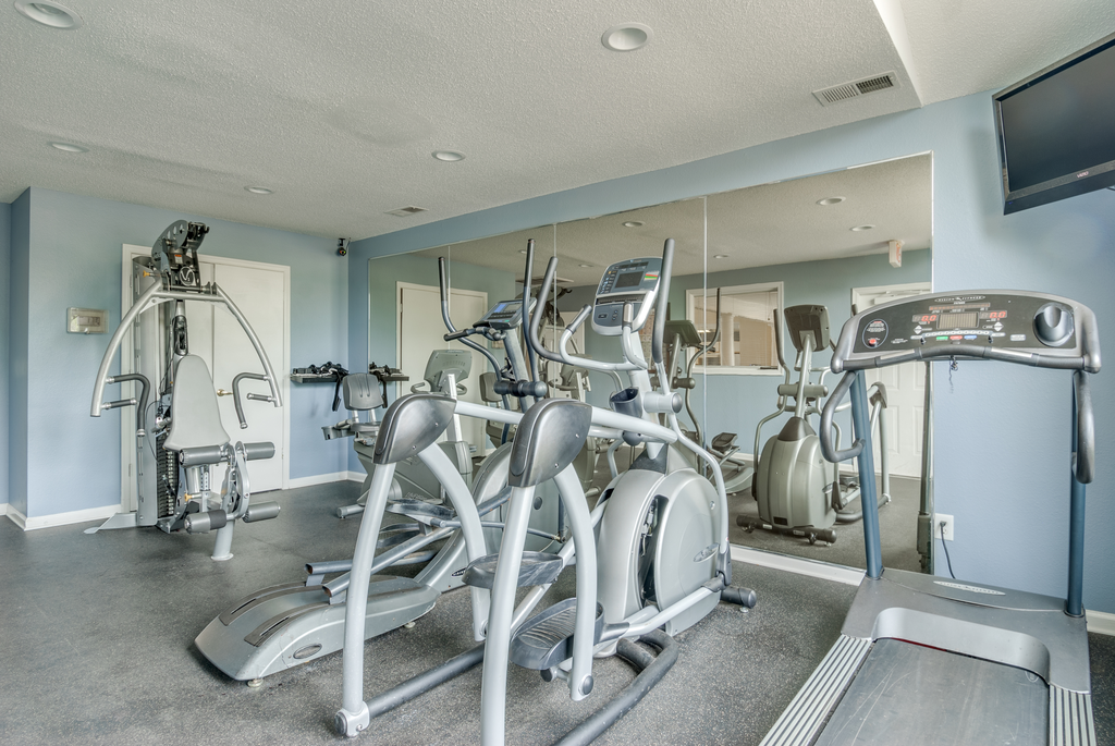 The community fitness center is equipped with a treadmill, two ellipticals and a weightlifting machine. One walls is made of mirrors and there is a TV mounted in the corner.