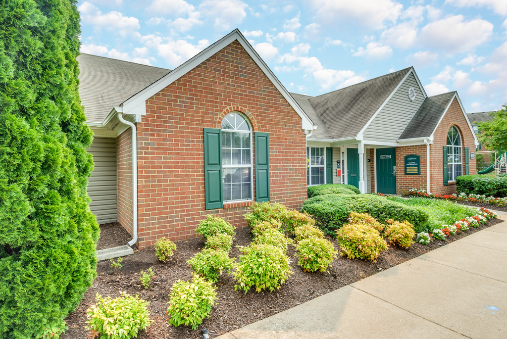 The Leasing Office building is mainly brick with a small amount of gray siding and forest green shutters and front door. There are flower beds on either side of the sidewalk leading to the front door.