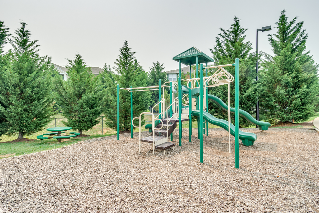 The community playground is surrounded by mulch and mature trees, and is located to the side of the clubhouse. The jungle gym is green and tan, has stairs leading to three slides and monkey bars.