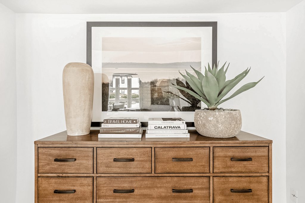 A wooden dresser with a framed picture on top, a vase, a book, and a plant at Paces Pointe Apartment Homes, Matthews, North Carolina