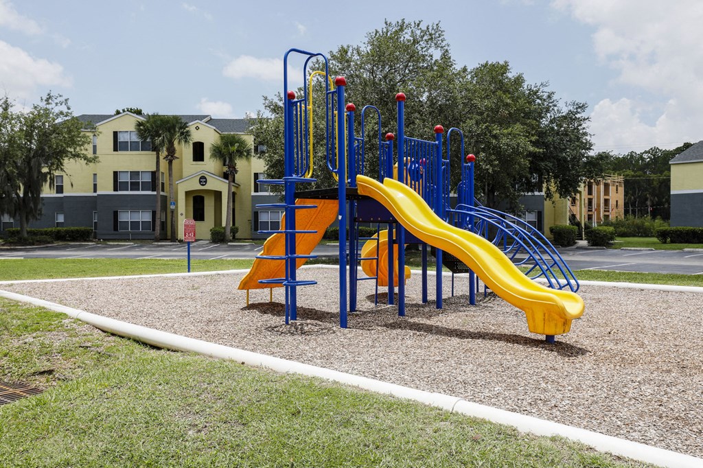 Playground jungle gym and playground slides in a bed of mulch with trees, and building exteriors in the background