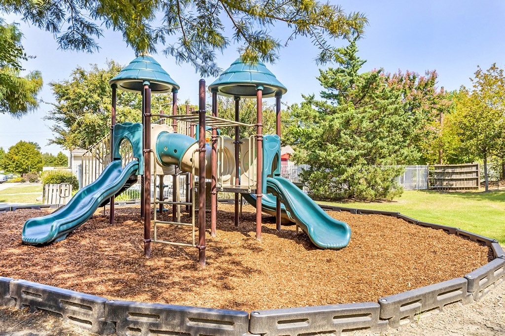 Playground jungle gym and playground slides in a bed of mulch with trees in the background