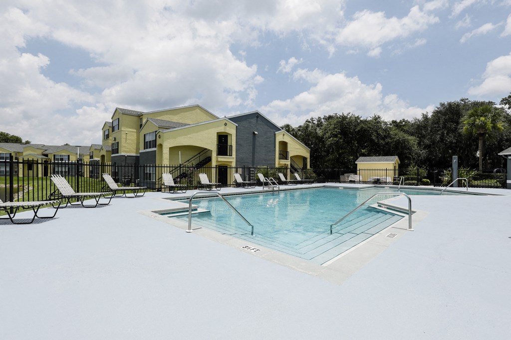 Community pool with sundeck, lounge chairs, surrounded by black metal fence with trees, and building exteriors in the background
