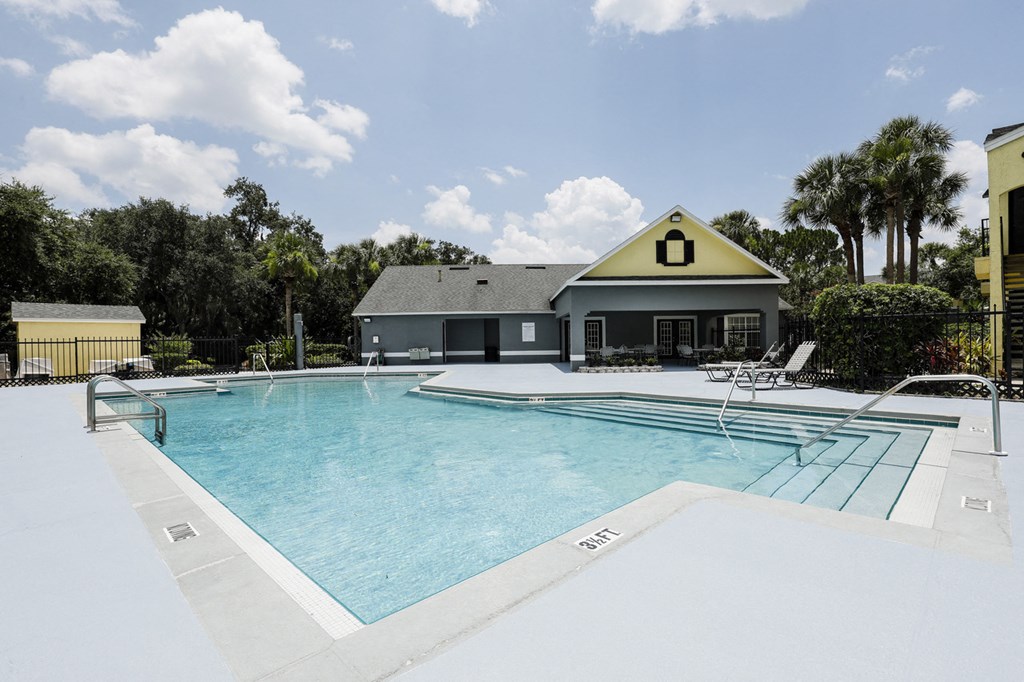 Community pool with sundeck, lounge chairs, surrounded by black metal fence with trees, and building exteriors in the background