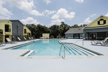 Community pool with sundeck, lounge chairs, surrounded by black metal fence with trees, and building exteriors in the background