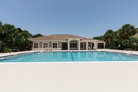 Community pool with sundeck, lounge chairs, surrounded by black metal fence with trees in the background