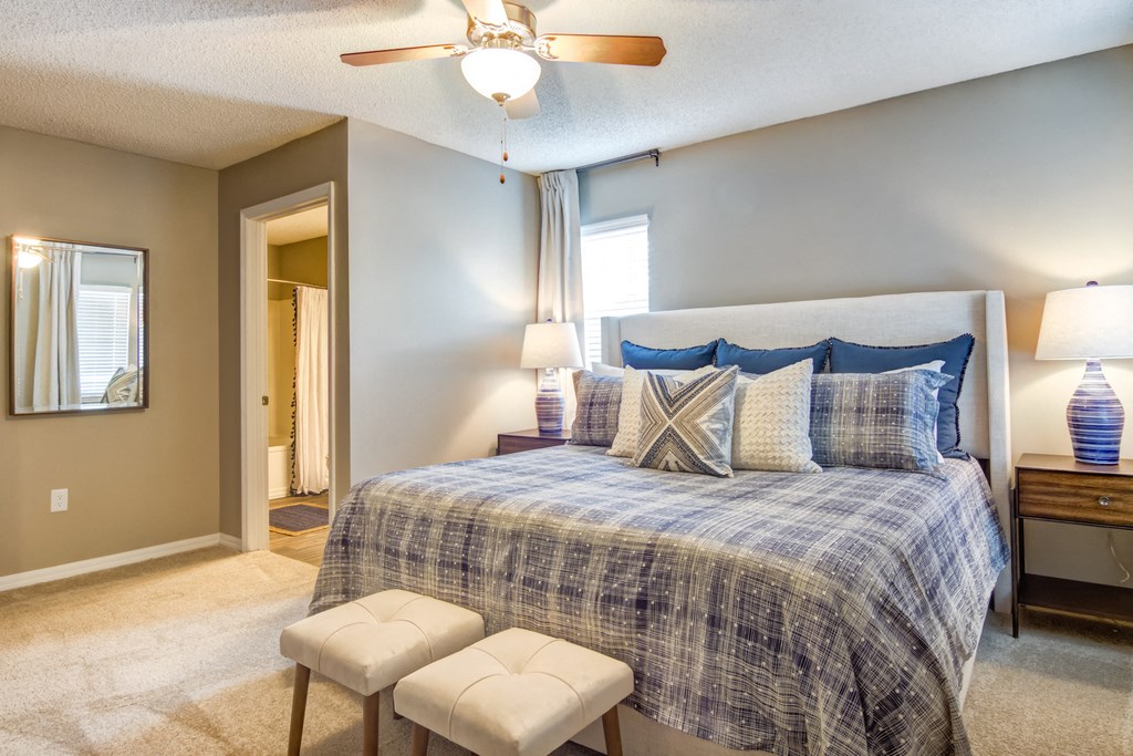 Staged bedroom with a window, gray walls, carpet, bed with seating in front, two nightstands with lamps, ceiling fan, decorative mirror and en-suite bathroom in background.