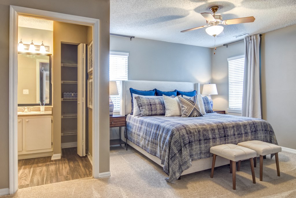 Staged bedroom with two windows, gray walls, carpet, bed with seating in front, two nightstands with lamps, ceiling fan, and entry way to en-suite bathroom in background.