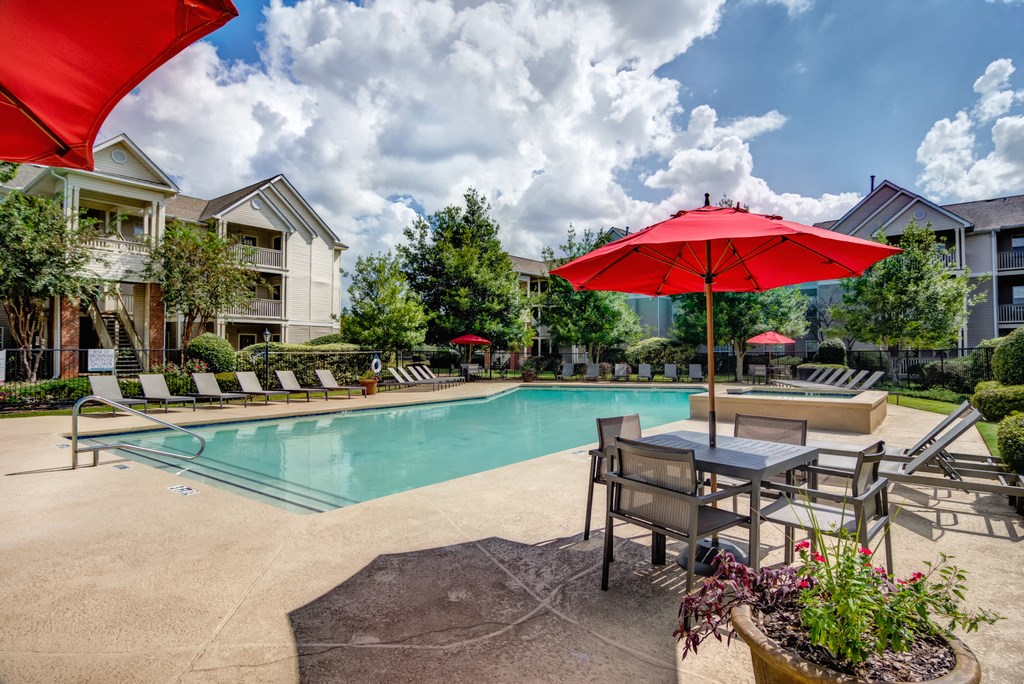 Swimming pool and sun deck with lounge chairs, tables with umbrellas, with native trees and building exteriors in the background.