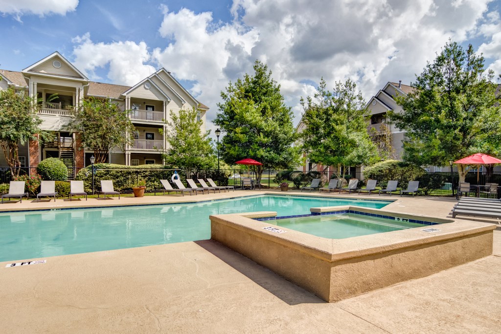 Swimming pool and spa, with sun deck with lounge chairs, tables with umbrellas, with native trees and building exteriors in the background.