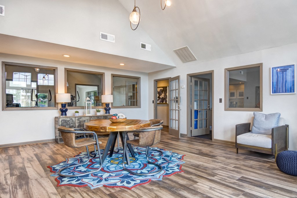Clubhouse interior with wood table and four chairs, a decorative chair with ottoman, and three interior widows looking into the Fitness Center.