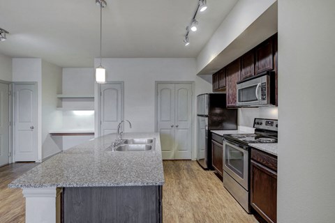 Kitchen with island, dark wood cabinetry, stainless appliances with wood style flooring, granite countertop, track lighting, and built-in desk in background