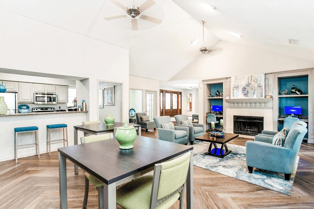 Clubhouse interior with high top tables and chairs, wood style flooring, fireplace with mantel, doors and windows, vaulted ceiling and ceiling fans, kitchen, and desks with computers and chairs
