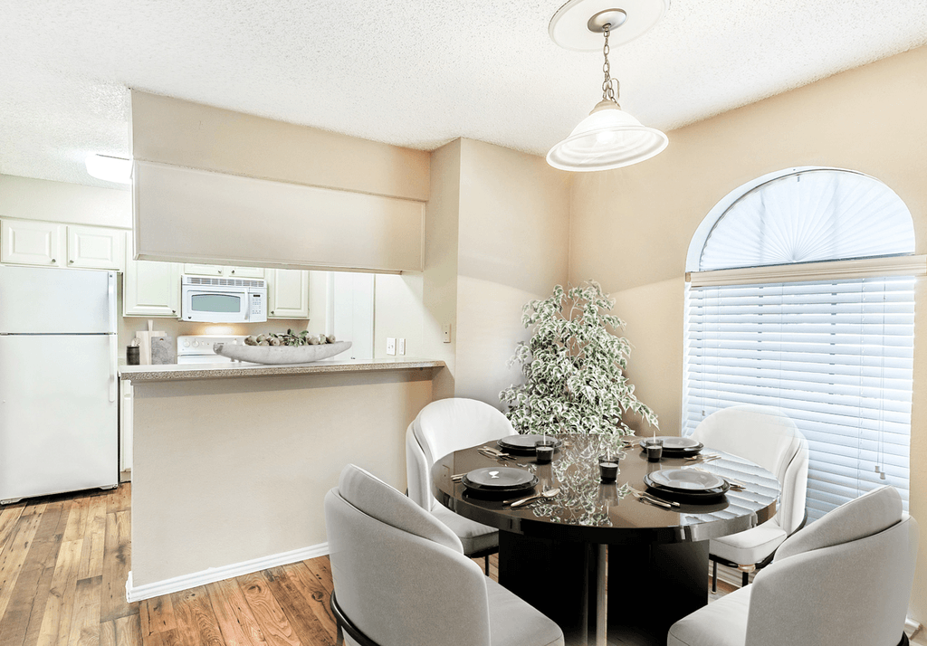 Virtually staged dining room with round table and chairs, wood style flooring, hanging light fixture, large window with blinds, and kitchen with breakfast bar in background