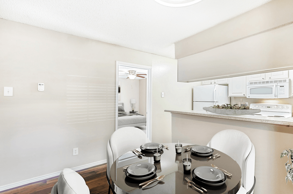 Virtually staged dining room with round table and chairs, wood style flooring, hanging light fixture, large window with blinds, and kitchen with breakfast bar in background