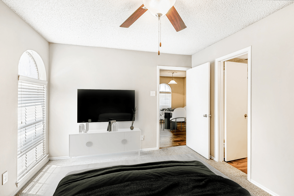 Virtually staged bedroom with carpet, accent rug, bed, ceiling fan, wall mounted television, tv stand, large window with blinds and en-suite bathroom
