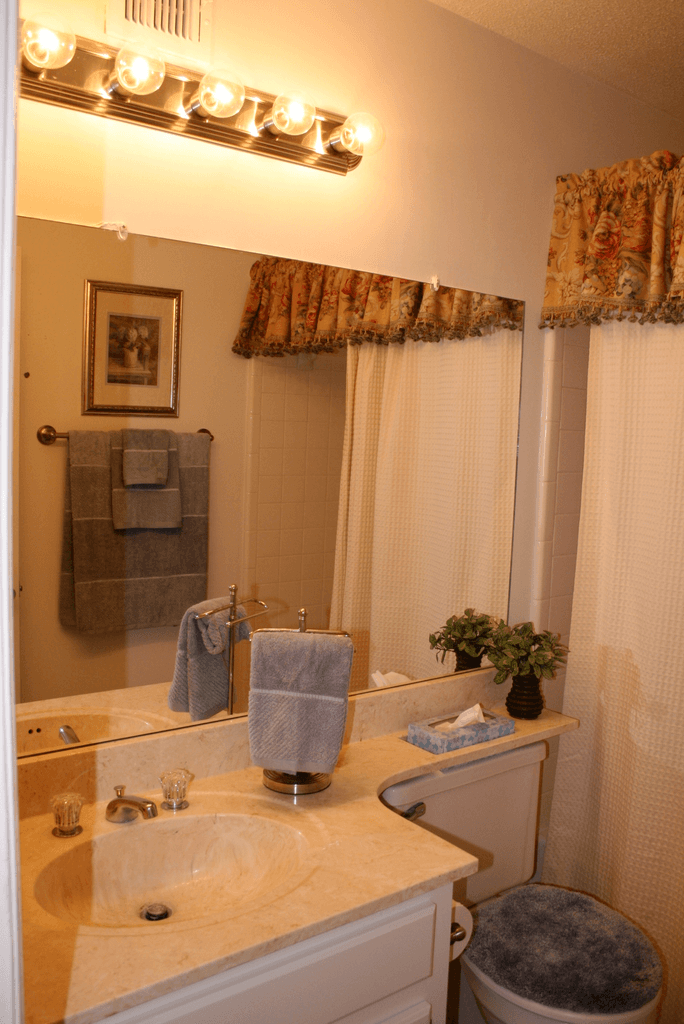 Staged bathroom with granite style countertops, white cabinets, large vanity mirror, shower and tub with white tile walls, white shower curtain, and gray towels.