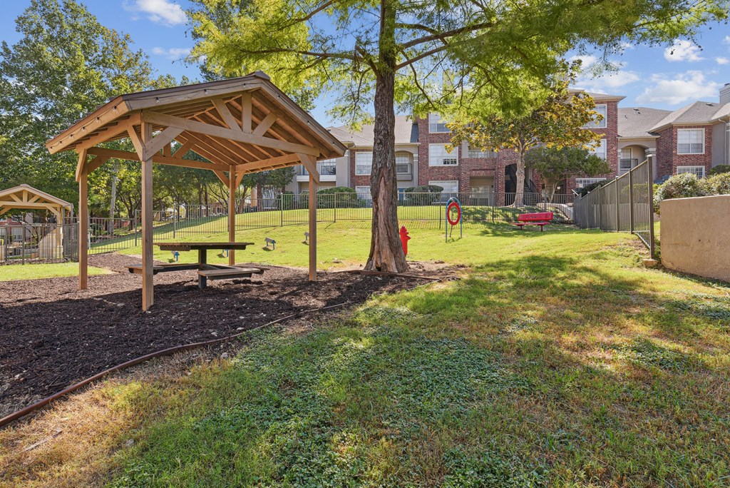 A wooden pavilion sits in a grassy area with a playground in the background.