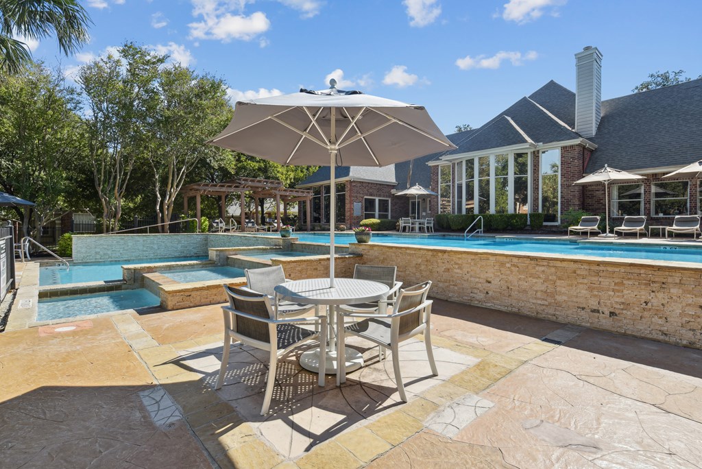 A patio with a table and chairs under an umbrella next to a pool.