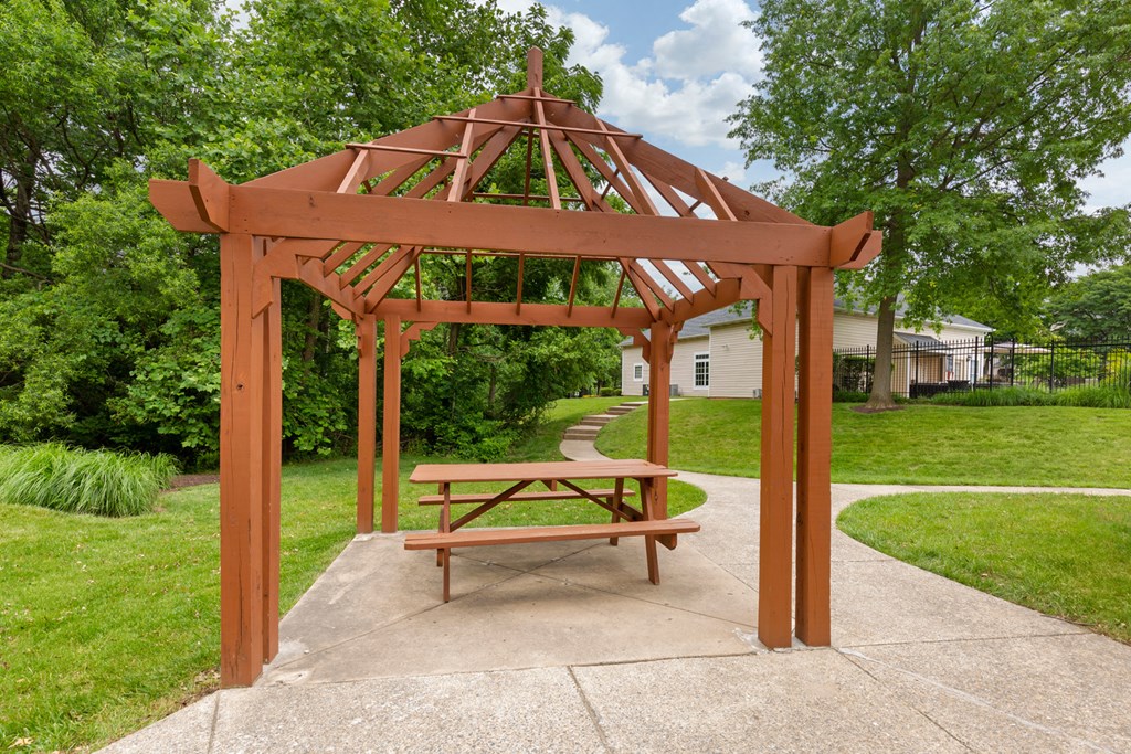 Wood gazebo with picnic table next to walkway surrounded by green grass and mature trees