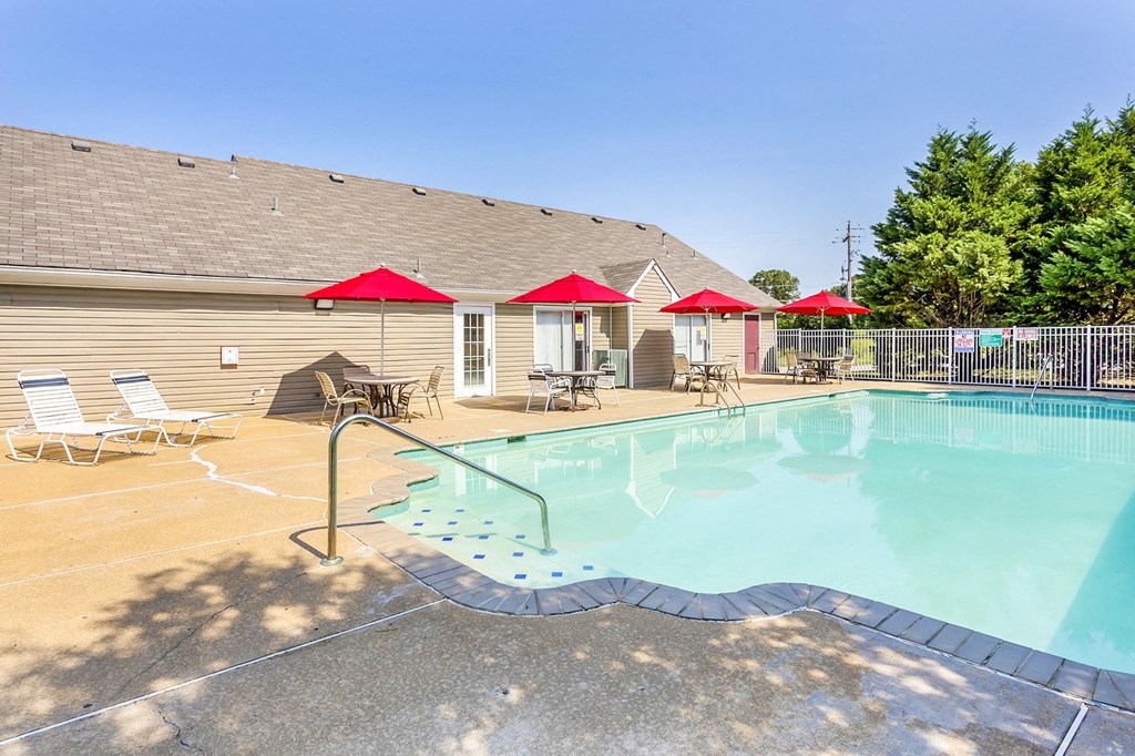 Community pool with sundeck, lounge chairs, surrounded by white metal fence with trees in the background