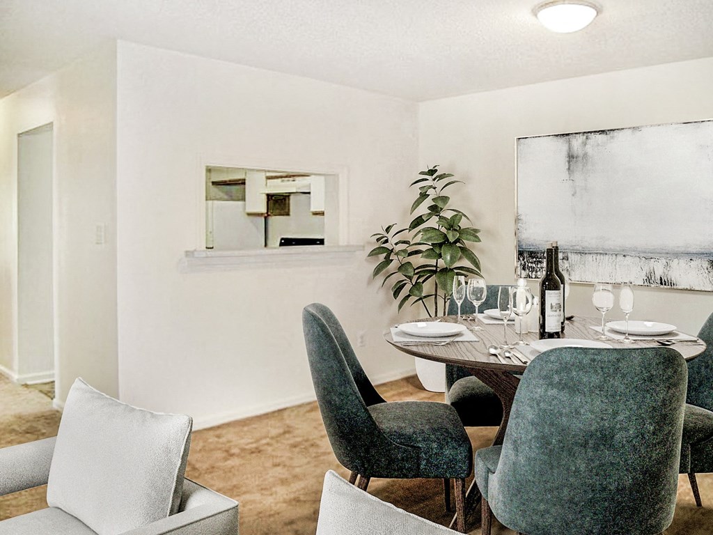 Carpeting dining area with white walls and view of kitchen in the background. Round dining table and chair set in the corner with art above the wall.