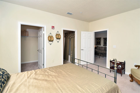 Decorated bedroom with tan carpet, beige walls, and ceilings. Bed lies on the left side of the room with a closet in the back