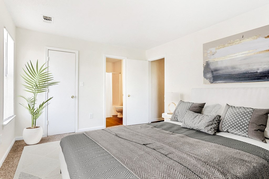 Staged bedroom with queen sized bed, art above bed, nightstand, large window in the background and view of bathroom and hall.