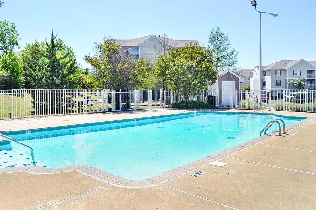 Community pool with sundeck, lounge chairs, surrounded by white metal fence with trees, and building exteriors in the background