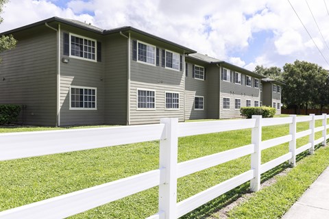 A white picket fence separates a grassy area from a building with multiple windows.