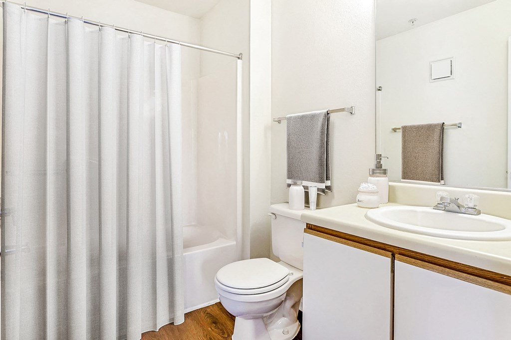 Staged bathroom with tiled bathtub shower combo. White counter tops, brown cabinets with a white faces, and wood style flooring. Grey towels hang on towel racks and lotions decorate the countertops.