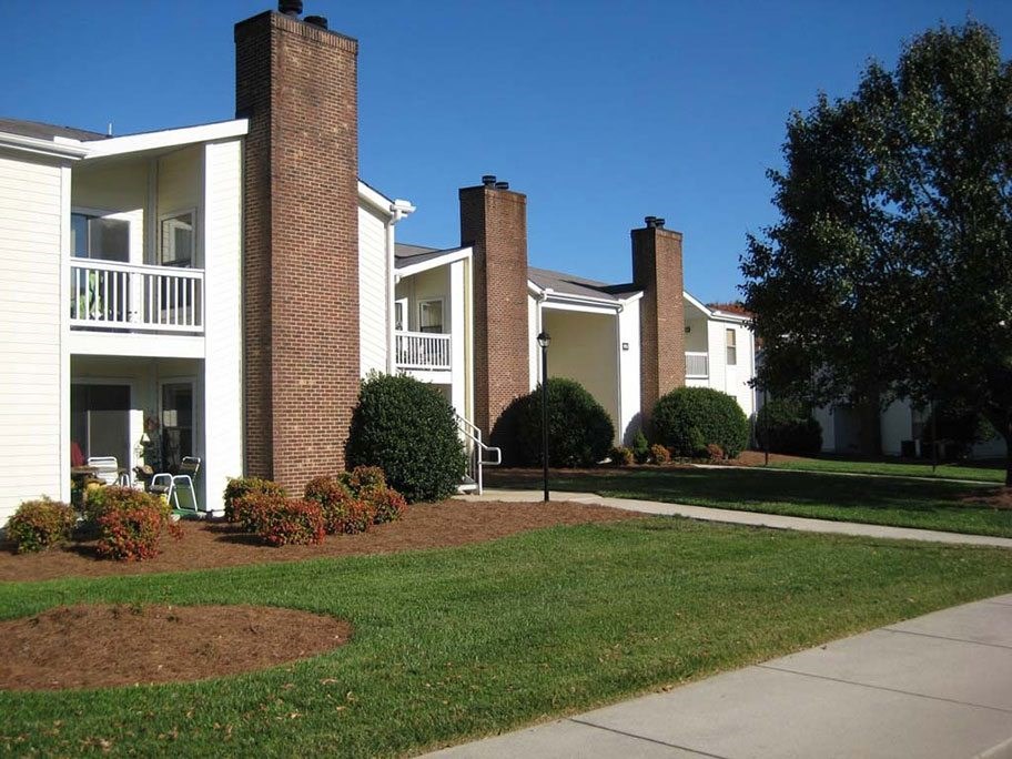 A white building with a brick chimney is surrounded by greenery at Fox Hollow Apartment Homes, North Carolina