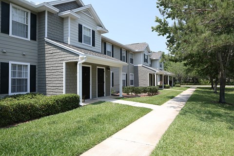 The resident building is a grey shingle sided building with white trimmed windows. In front of the entrance is a concrete walkway that extends to both sides to a sidewalk. The landscaping on either side of the entrance is lush green grassy plants, brown mulch, and large green bushes.