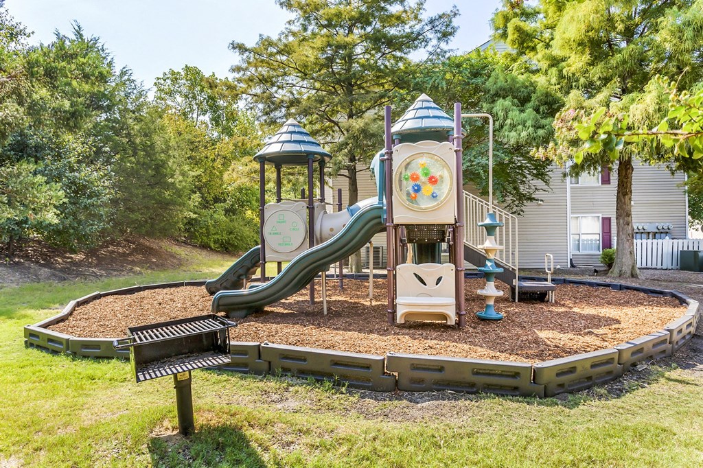 Playground jungle gym and playground slides in a bed of mulch with trees in the background and a grill in front of it